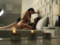 PaolaYKatty with long black hair, naked, on top of another PaolaYKatty with black mesh top, on bed with tufted headboard, candles in foreground, intimate, soft lighting.