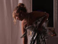 RosaVance bends forward, wearing a shiny, sequined dress that's slipping off her tits, with blonde hair in a messy bun, and a pearl necklace, in dim, moody lighting.
