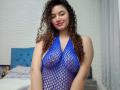 SanetPerlas with curly brown hair, blue fishnet dress showing her tits, pink lipstick, and a confident smile, standing in a bedroom with a blue headboard.