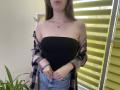 Nesie with bare tits, black strapless top, plaid shirt off shoulders, blue jeans, long brown hair, necklace, standing in front of yellow blinds, green plant in bottom left.