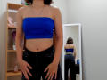Steisii with blue tube top, showing her bare tits, black jeans, and tattooed arm, standing in front of mirror reflecting her ass. Room with shelf and stuffed pink bear.
