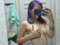 Topless MathaDernier with purple hair, covering her tits with one hand, taking a mirror selfie with a blue phone, showing a tattoo on her right arm.