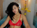 AriaVega with curly black hair, wearing a red lace bra, showing her tits, sitting in a chair, yellow flowers in the background, and a lamp to the right.