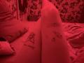 DarylSanks' legs are spread on a bed, showing tattoos on their inner thighs—pizza with text and a donut on the right leg, and a small heart on the left. Red lighting.