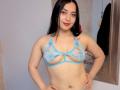 MelaniCoox with pale skin, black hair, and small tattoos, wearing a blue lace bralette that reveals her tits, standing with hands on hips, smiling slightly.