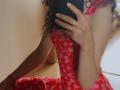 SweetClaraa with curly brown hair, wearing a red floral dress, taking a mirror selfie with her phone, showing side boob, ring on her finger, kneeling on wooden floor.