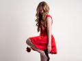 AkilahGhil, red dress, long wavy blonde hair, holding red rose, black high heels, right leg raised, looking down, white wall background, bracelet on right wrist.