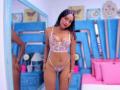 CassidyWinslow stands in a blue room wearing a sheer white lace bra and thong, showing her small tits and pussy, with curly dark hair and a slight smile.