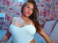 BelletinaXx with medium brown skin, long brown hair, pink lipstick, white ribbed tank top, showing her large tits and tattoos, smiling slightly, standing in a room.