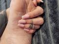 ElixirShanna's hand holds another hand, showing tattoo on ring finger, pink nails, and slightly wrinkled skin. She's wearing a dark, textured shirt.
