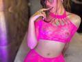 AprilBoo en lingerie en dentelle rose, montrant ses petites seins et son ventre plat, avec son doigt près de sa bouche, portant des lunettes, sur une image légèrement floue..