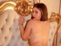 MelanyRios with a bob, wearing a white string bikini, has her back to the camera, looking over her shoulder, with gold ornate headboard and tufted beige upholstery.
