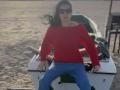 Nalini, con i capelli neri e gli occhiali da sole, indossa una maglietta rossa a spalle scoperte e pantaloni blu, seduta su una piccola barca sulla spiaggia, con le gambe divaricate. Barche sullo sfondo..