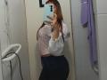 AntoniaBrunni with long red hair, wearing a pink floral top, white ruffled sleeves, and black jeans, taking a mirror selfie in a bathroom, showing her ass.