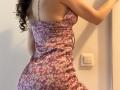SweetClaraa wearing a pink floral dress, showing her back and butt, with curly dark hair, standing near a white wall and light switch.