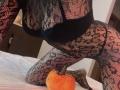 Euphorias is on all fours, wearing black lace bodysuit, showing her tits and ass. Small orange pumpkin is near her knee on white bed.