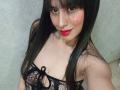 MadissonFontaine with long black hair, red lipstick, and a black lace top showing her tits, smiling at the camera. She has a small mole on her right cheek.