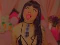 AnnieGrey with black hair, plaid tie, and choker, sticking out her tongue, tattoos on arms, pink makeup, wearing strappy black top, pink and pinkish background.