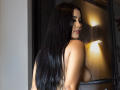 VanesaNova with long black hair, looking over her shoulder, showing her tits, standing near a window with warm light. Her skin is tan, and she's smiling slightly.