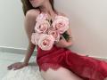 StellaNoir, wearing a red lace lingerie dress, sits on a white rug, holding a bouquet of pink roses, covering her tits. Her red dress contrasts with her pale skin.