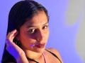 MelodyRoss with dark hair, brown skin, and red lipstick, touching her ear with a hand, wearing a black top and pink necklace, against a blue background.