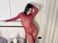 ZammySanz in a red lace bodysuit, showing off her curvy ass and small tits, leaning against a wall, smiling, with one hand on her head, in a bedroom.