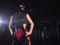 SofiBrandford in een zwart leren outfit met een rood drakenmotief, een masker dat haar gezicht bedekt, handen op de heupen, staand in een zwak verlichte kamer, een spotlicht van links, die haar rug en zijkant laat zien..