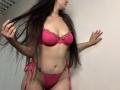 Tisia with long brown hair wearing a pink lace bra and matching thong, holding her hair, showing her tits and pussy, against a white wall.