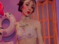 LaurenEvie69 with pale skin, short black and white hair, red lipstick, white lace bralette, tattoo on chest, pink bow, silver choker, pink donut inflatable on left.