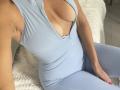 SandraSweet in a light blue, zip-up, form-fitting, sleeveless top showing her tits with a deep V-neck, sitting with her hand on her leg, wearing matching pants.