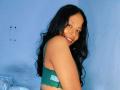 AshleyGattes with long black wavy hair, smiling, wearing a green lace bra, showing her small, cute ass, and small, perky tits, with hoop earrings, in a blue-walled room.