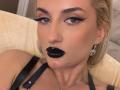 MademoiselleBella has black glossy lipstick, thick black eyeliner, and black mascara on blue eyes, wearing black strappy top and silver earrings, with blonde hair and tan skin.