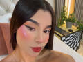 LunaVali with shiny pink blush, red lips, black eyeliner, and long dark hair, looking down, has cute face and small nose. Bedroom setting with potted plant and laptop.