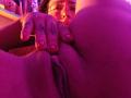 MilaThorne's face with glasses, her hand spreading her ass, revealing her pussy. Her fingers have tattoos and painted nails, in pink-purple lighting.