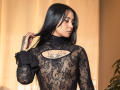GabyMathew with long black hair, wearing a sheer black lace top, showing a tattoo on her chest, touching her hair, looking to the side, in a warm-lit room.
