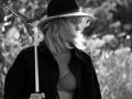Black-and-white photo of EolantaMarina with blonde hair, wearing a hat, dark coat, and bra, holding a net, looking down with a slight smile. Her tits are visible.
