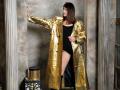 MiraMixi with shiny gold coat, black one-piece, and black boots, standing, left arm on wall, right hand on hip, looking sideways, in a gritty, industrial setting.