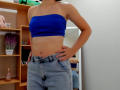 Steisii with blue tube top, showing bare midriff, hands on jeans, mirror reflecting her back, slightly visible tits, small waist, tan skin, casual room, bottle on shelf.