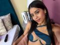LaurentRay with long black hair, brown eyes, and a nose ring, wearing a blue cut-out bikini top, smiling at the camera, with a black vibrator on the bed.