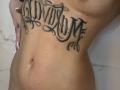 AuraXo's bare torso with a black cursive tattoo across the chest, showing small breasts and a slightly visible belly button. Her hand with long nails is on the left.