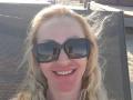 MysteryLaddy with blonde hair, wearing black sunglasses, smiling at the camera, lying on a mesh chair, sunlit face, visible teeth, and a slight sunburn on the nose.