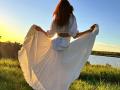 Il vestito bianco di MiraGi svolazza mentre lo tiene aperto, mostrando il suo sedere in un prato soleggiato, con un fiume sullo sfondo e un cielo azzurro limpido..