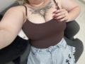 DiLaria with large tits, brown tank top, red nails, sun tattoo on chest, holding top, wearing frayed jeans, sitting on black chair, blonde hair, hand on chest.