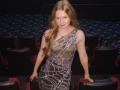 AkilahGhil with long blonde hair, wearing a sheer, silver-sequined dress, exposing her small tits and nipples, standing in a theater, leaning on a black seat.