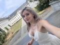 EliseMonet, avec ses cheveux blonds platine, souriante, portant une robe blanche avec un décolleté, prenant un selfie, montrant ses seins, dans un cadre extérieur ensoleillé avec un bâtiment historique..