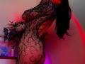 Tsukerberg with long black hair, wearing a black lace bodysuit, showcasing her ass and back, in a red and purple lit room, holding a bouquet of flowers.