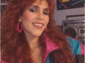 AntoniaBrunni with red curly hair, pink earrings, and a colorful jacket, smiling with white teeth, wearing makeup, looking cute and sexy, 80s vibe, boop-boop-boop.