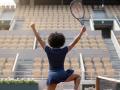 RoldanStefy, with curly black hair, wearing a navy blue tennis dress, jumps with arms raised, holding a tennis racket, standing in front of empty stadium seats.