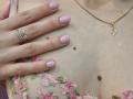 MilaYanis' pale skin, small brown mole, pink nail polish, gold necklace with flower charm, and pink floral lace bra, left hand with silver ring, touching her chest.