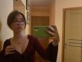 LeScharwath with glasses, brown hair in a bun, red dress, showing cleavage, holding green phone, ring on finger, looking at mirror, wooden door and wall in background.